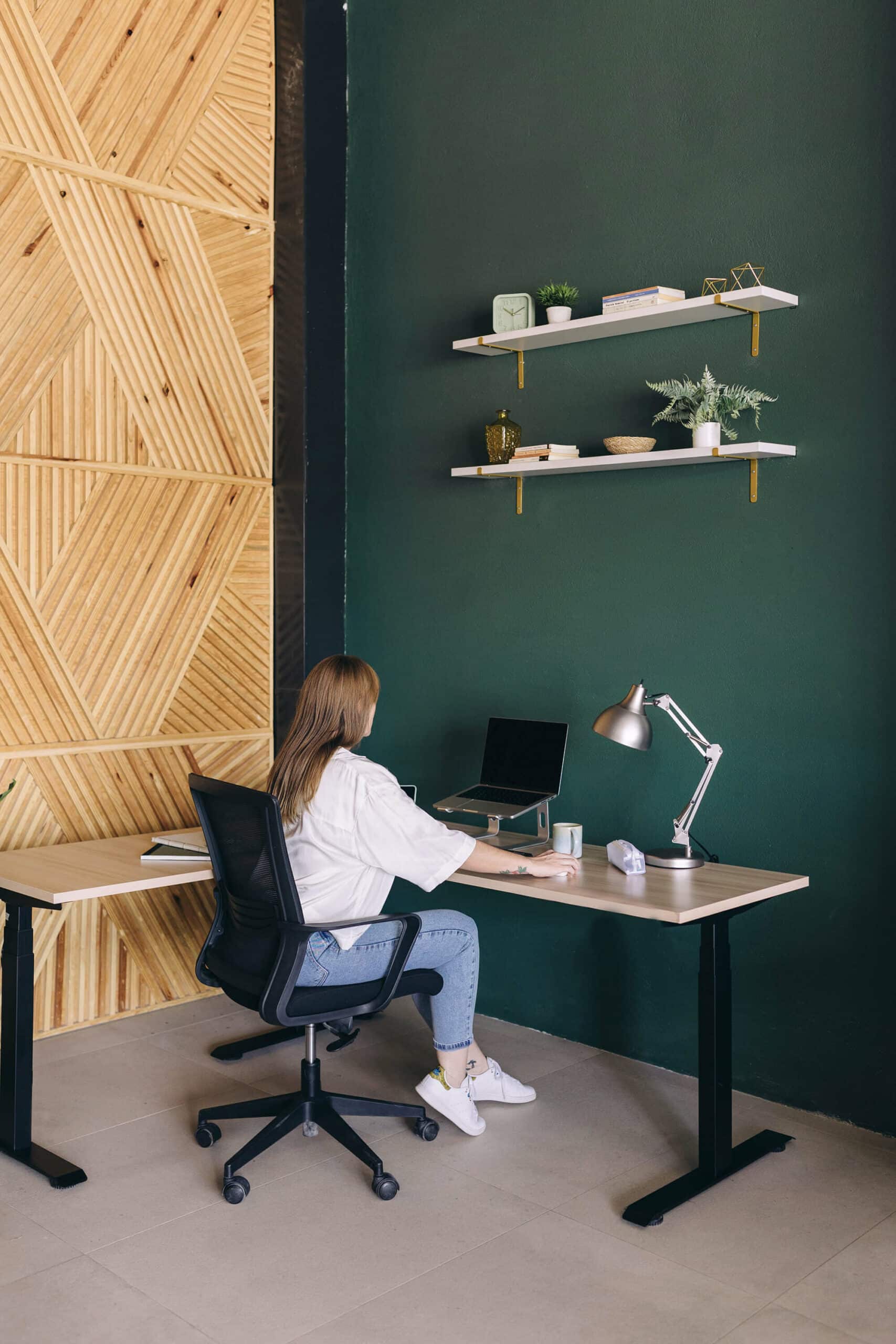 A woman sits at a modern wooden desk facing a wall painted green, working on a laptop which is elevated on a stand. The office has a stylish design with decorative shelves above the desk, an adjustable desk lamp, and a herringbone-patterned wood panel on the adjacent wall.