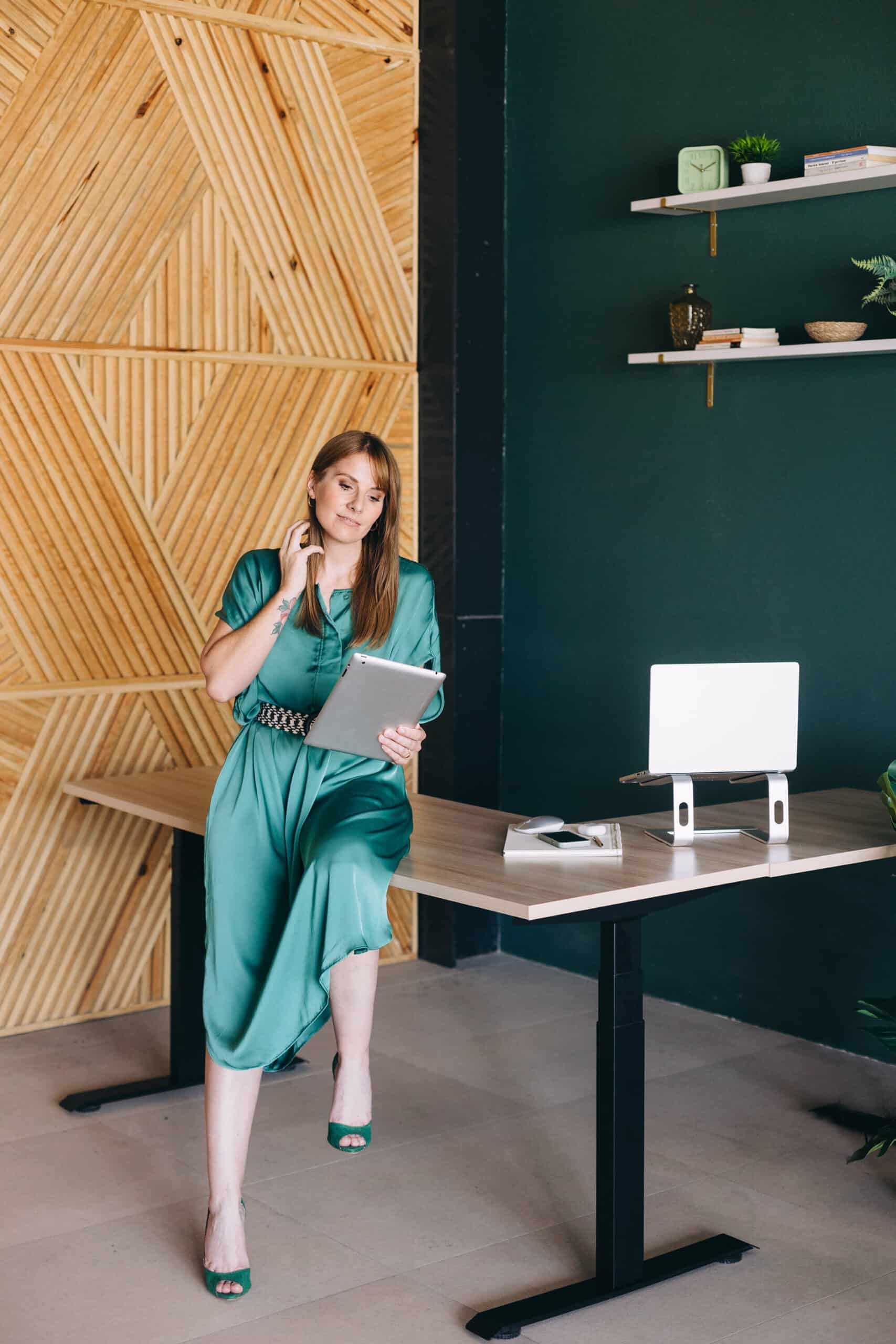 A woman in a green dress and matching heels is sitting casually on a desk, holding a tablet and talking on the phone in a modern office with wooden geometric wall decor and minimalistic shelves.