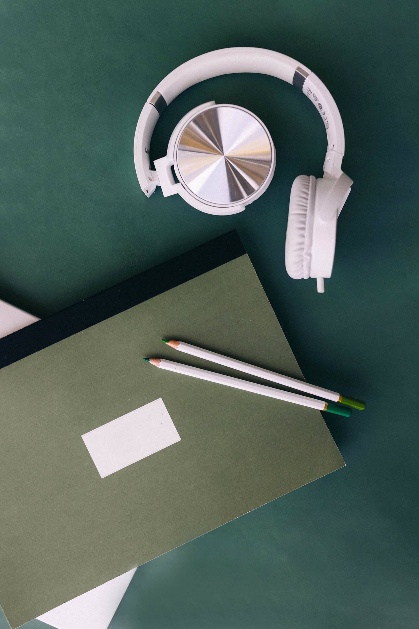 A pair of white over-ear headphones laid on a green surface next to a dark green notebook with a blank white rectangular label attached to it and two pencils on top.