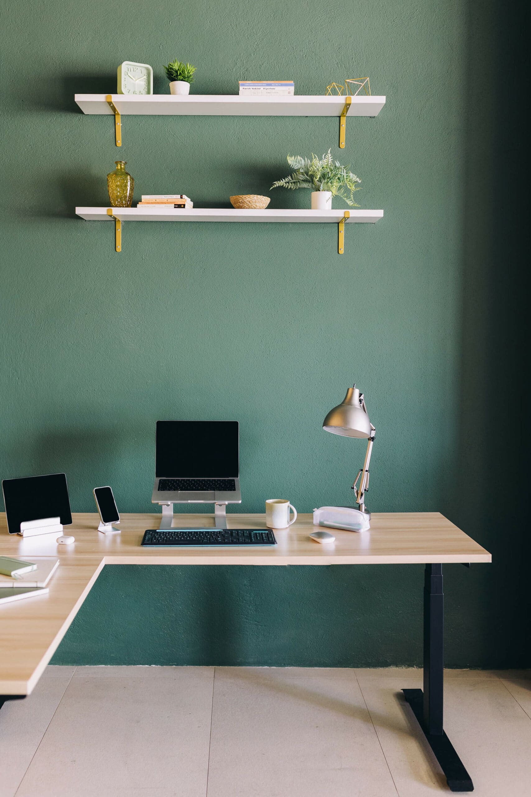 A minimalist home office setup with a light wooden desk against a green wall, equipped with a laptop raised on a stand, a tablet, a smartphone, a desk lamp, a cup, and some stationery. Two white shelves with decorative items and plants are mounted above the desk.