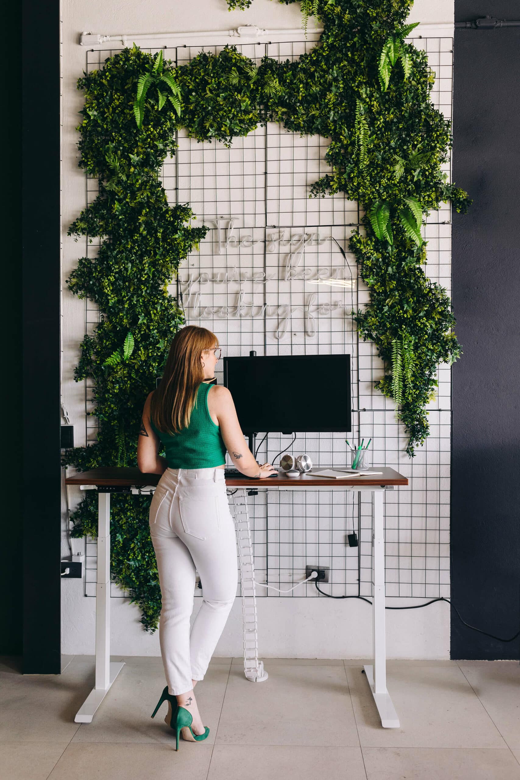 A woman in a green sleeveless top and white pants stands at a standing desk with a computer monitor, facing a grid wall with lush green foliage. She is viewed from the side and appears to be working or studying.