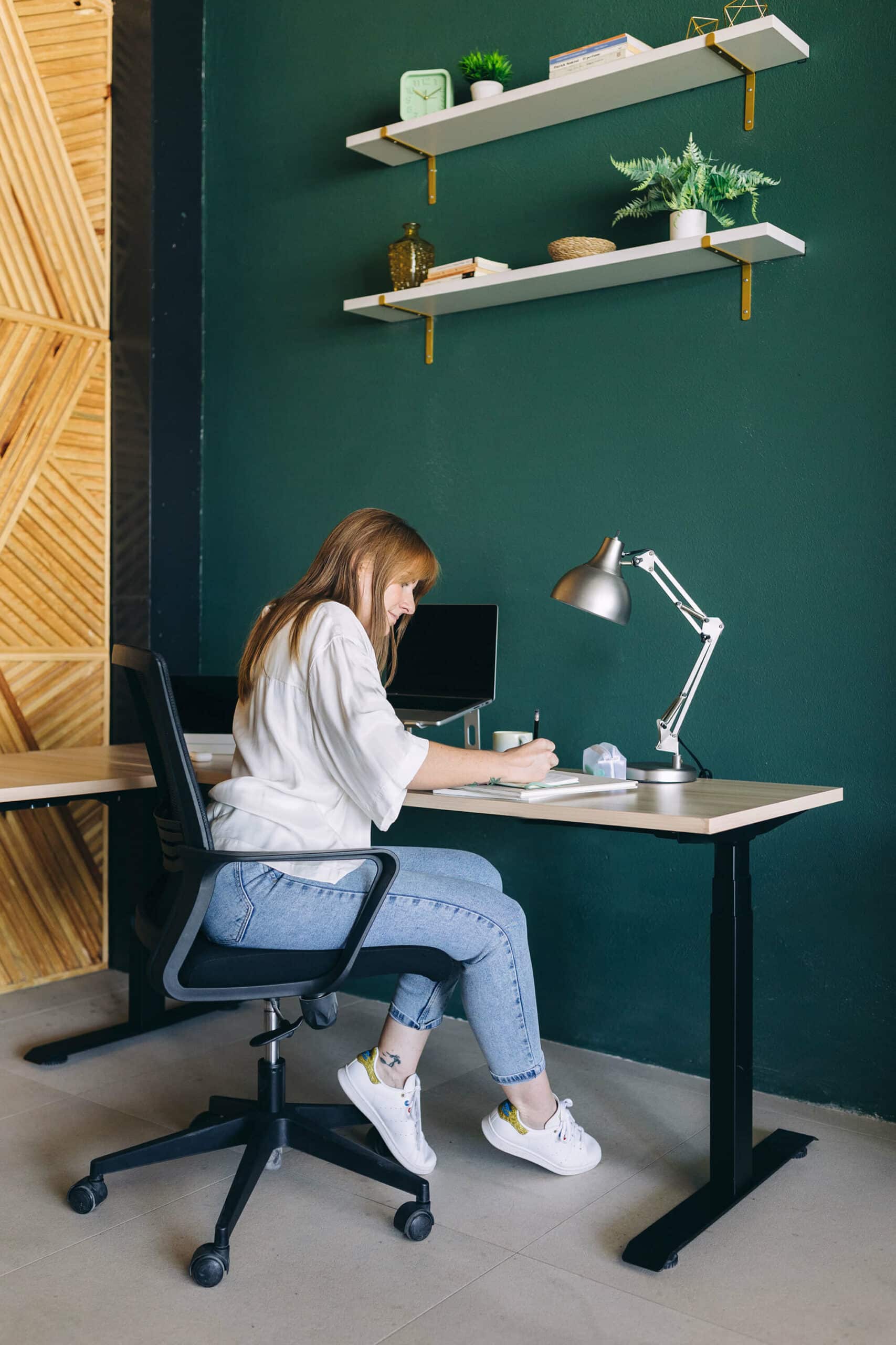 A woman sits at a desk in a modern home office, working on a laptop. The room features a dark green wall, floating shelves with plants and decorative items, and a wooden herringbone pattern on an adjacent wall. The woman is wearing a white shirt, blue jeans, and white sneakers, focusing on her notebook with a desk lamp beside her.