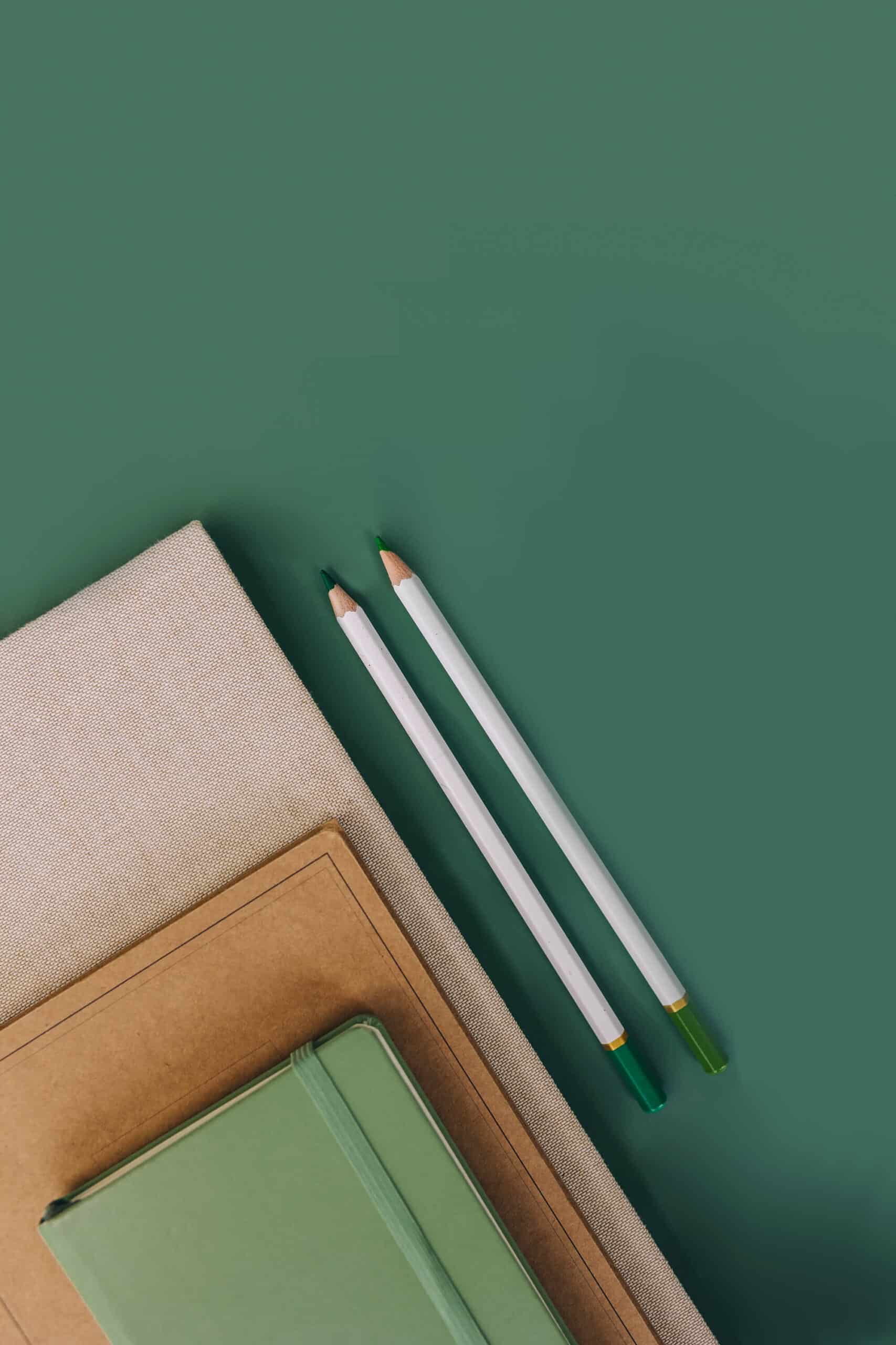 Two green-tipped white pencils placed on a green backdrop next to a stack of notebooks with a brown top notebook and a green one beneath.
