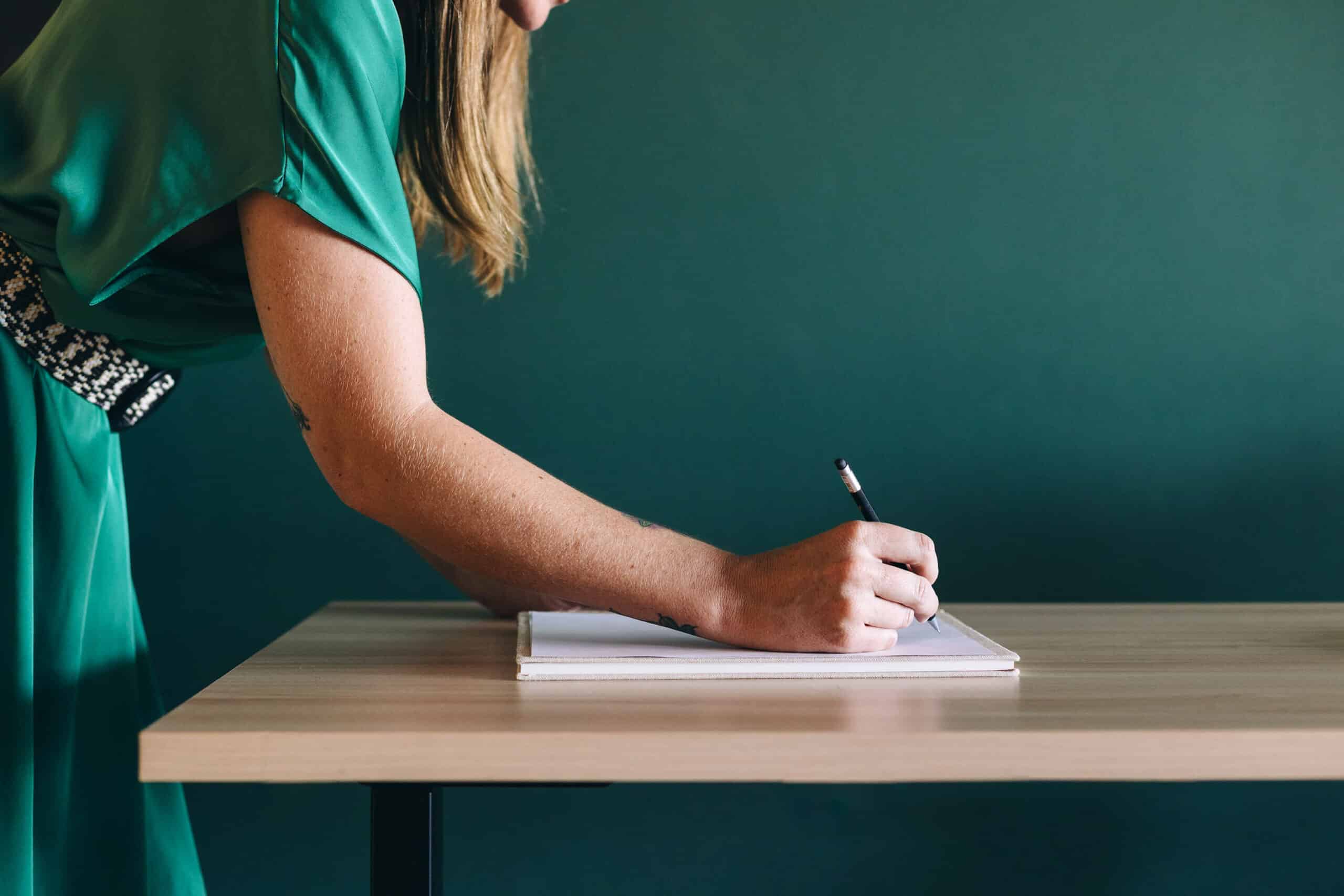 A person in a teal dress is writing in a notebook on a wooden desk against a green background. Only the person's right arm and part of their upper body are visible in the frame.