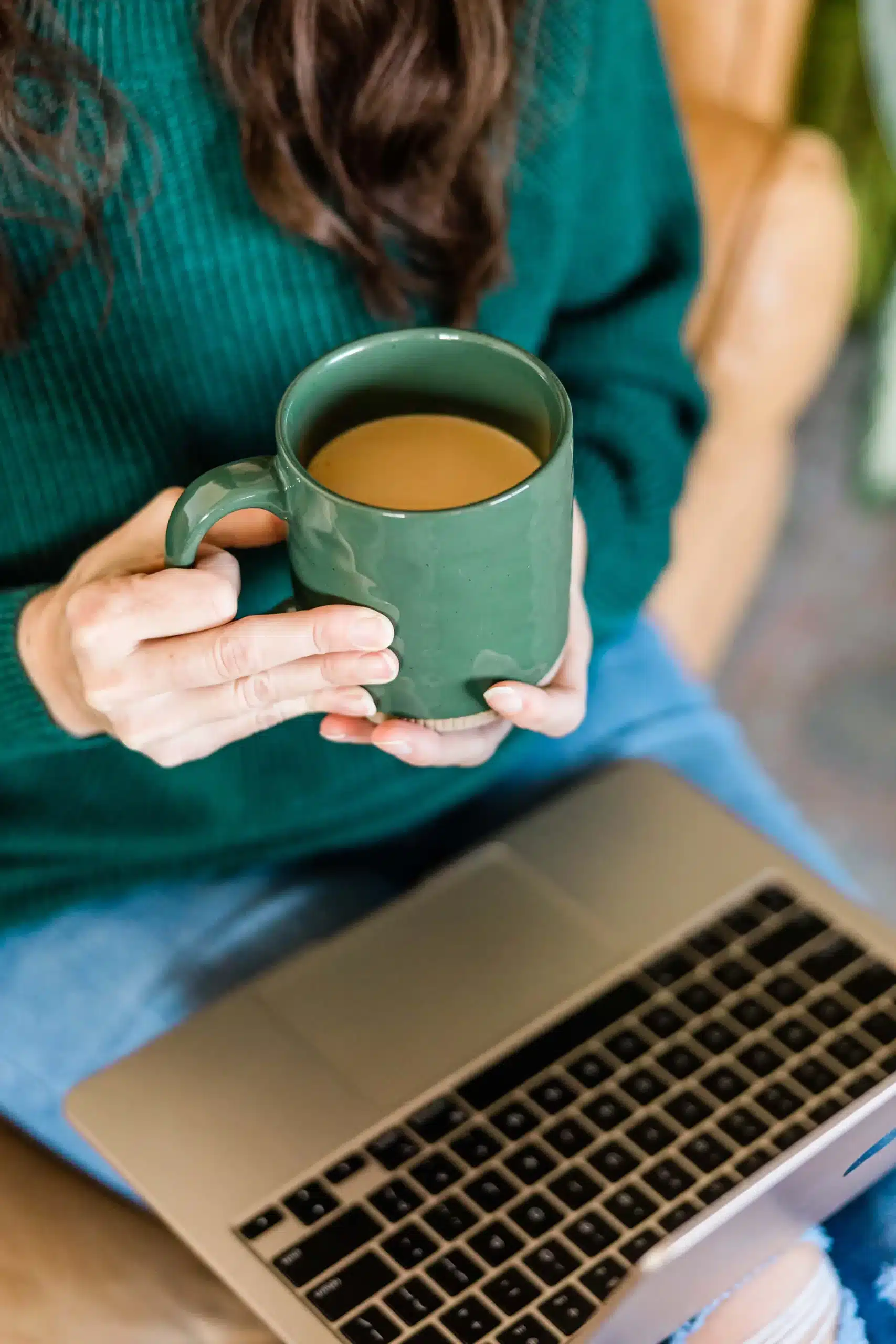 A person seated with a laptop on their lap holding a green mug with a beverage, wearing a green sweater, with only their torso and arms visible.